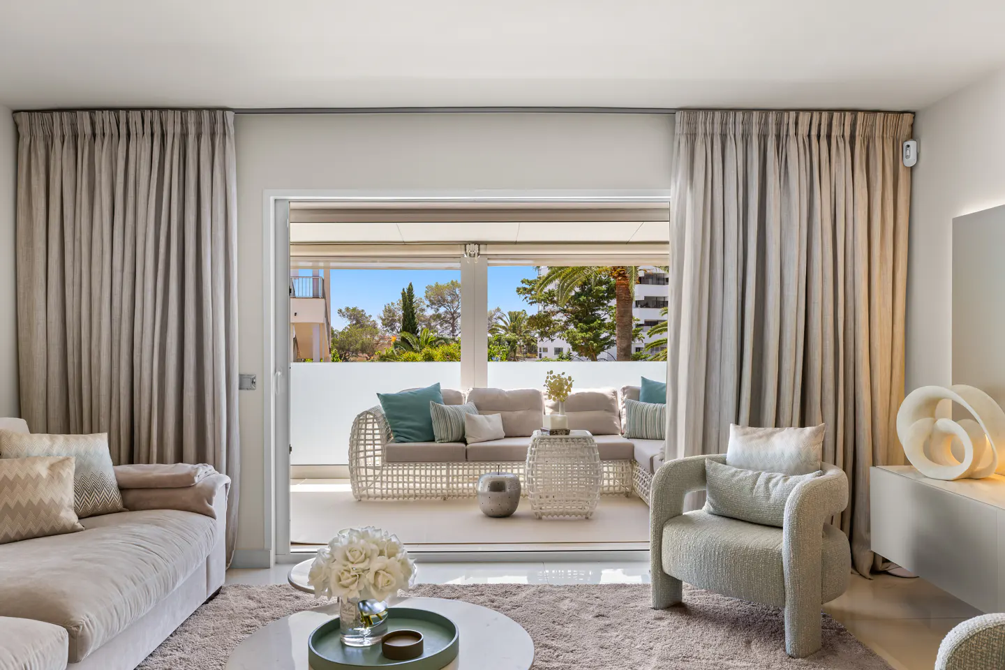 Living room with beige sofa, chair, and rug. Balcony with white wicker furniture and blue pillows visible through open sliding glass doors.