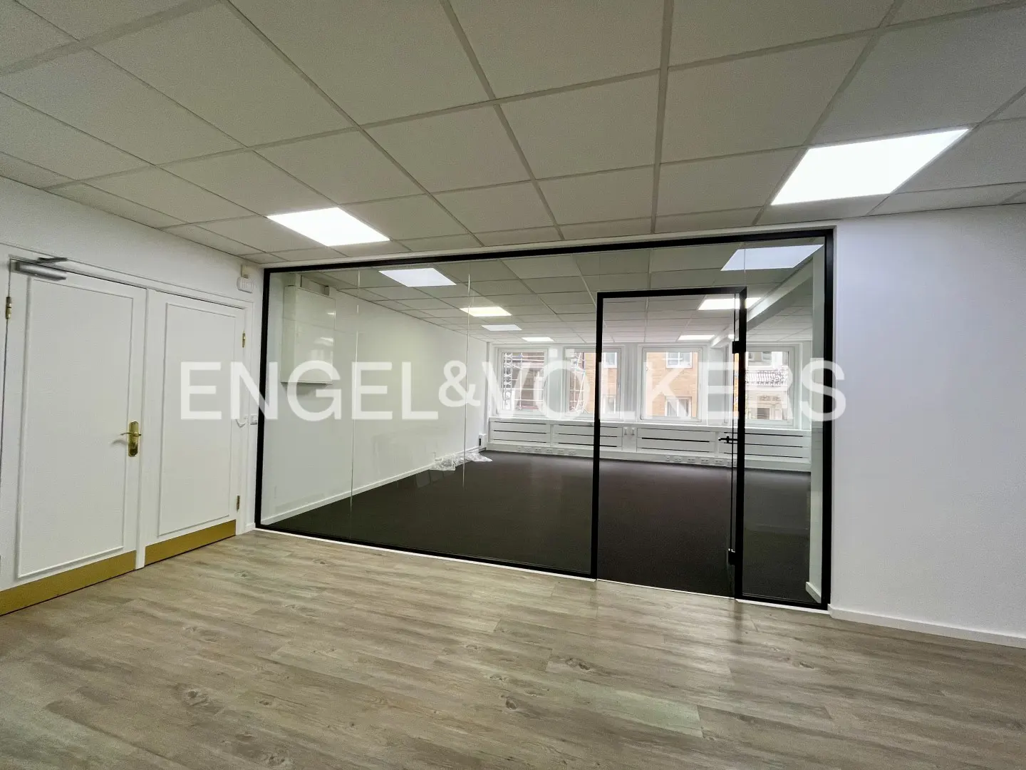 Empty office space with wood-look floors, white walls, and a glass-walled conference room with black trim. Two white doors are on the left.