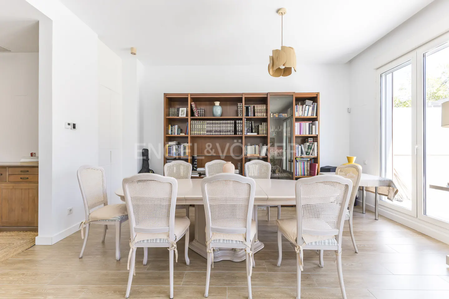 Bright dining room with a long table, white chairs, and a large bookshelf filled with books. A modern light fixture hangs above the table.