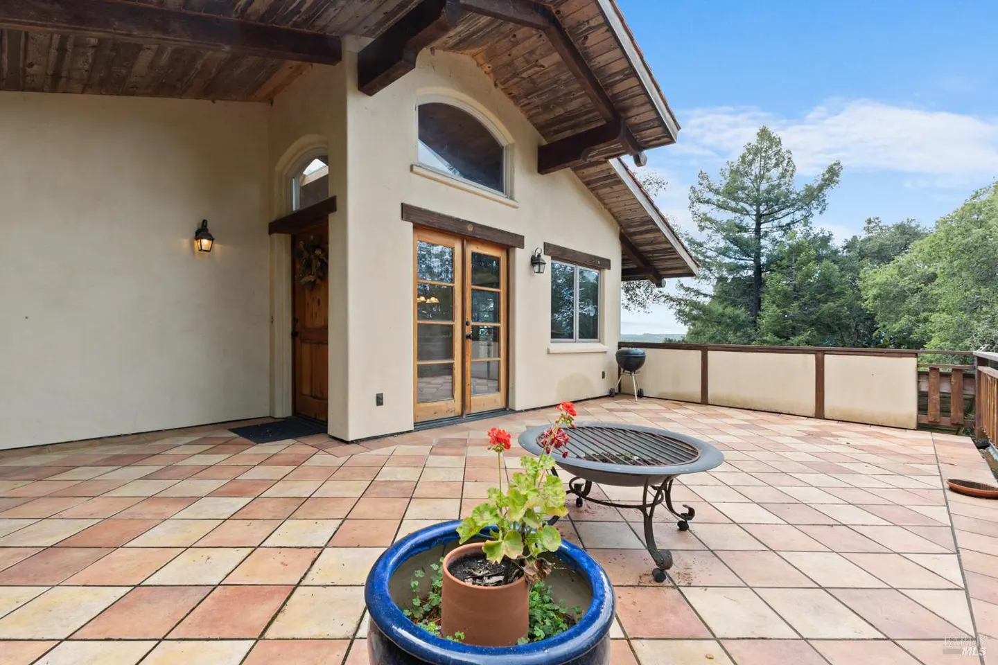 Exterior view of a beige house with a tiled patio, a fire pit, and a blue flower pot. Trees and blue sky are in the background.