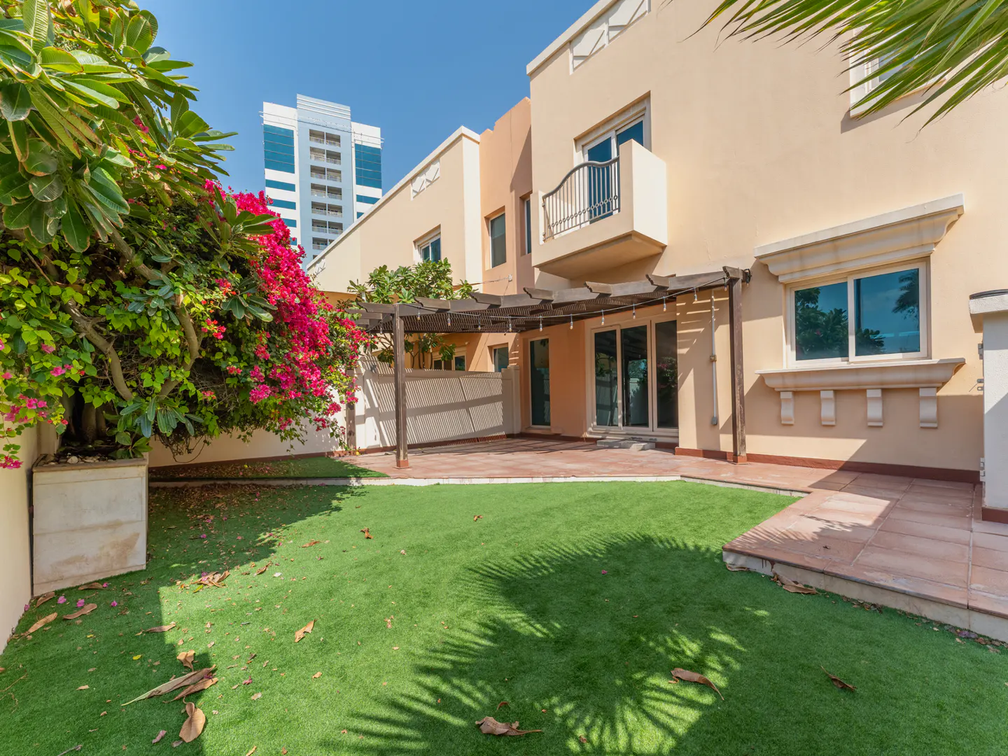 Backyard view of a two-story beige house with a green lawn, a patio with a pergola, and pink bougainvillea. A tall building is visible in the background.