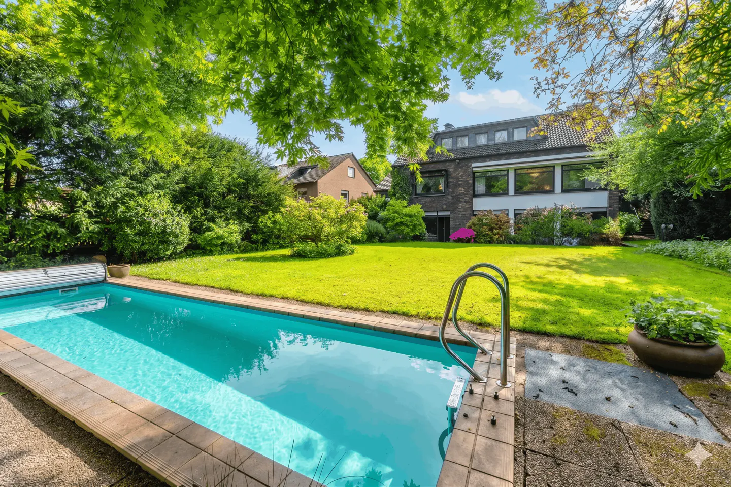 A backyard swimming pool with turquoise water, surrounded by green grass and trees. A house is visible in the background.