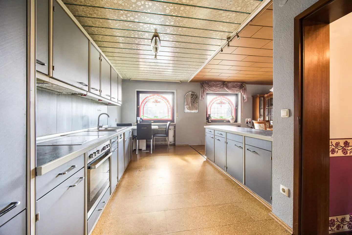 A kitchen with gray cabinets, a speckled ceiling, and two arched windows. A table and chairs are visible in the background.