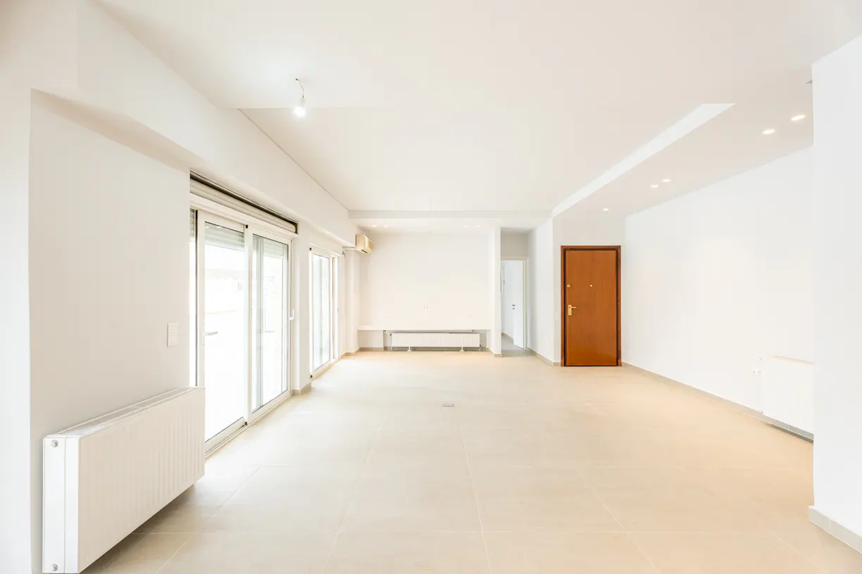 Bright, empty living room with white walls, tiled floors, a brown wooden door, and large sliding glass doors.