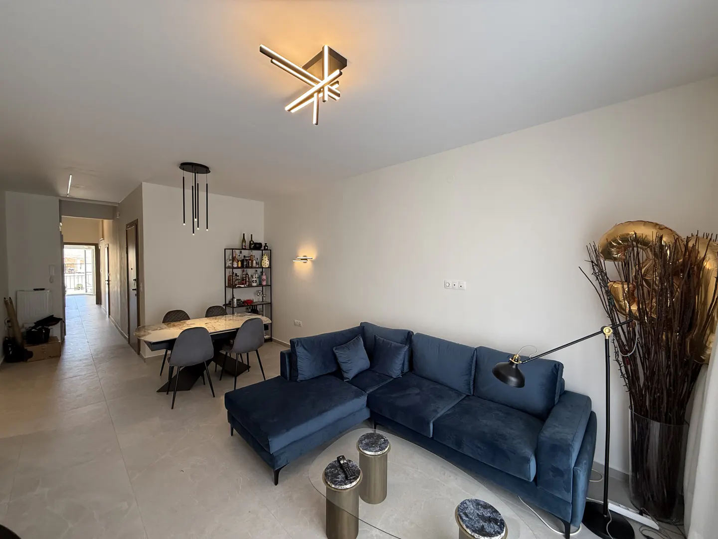 Bright living room with a blue sectional sofa, marble table, and modern lighting fixtures. A bar cart and gold balloons add a touch of elegance.