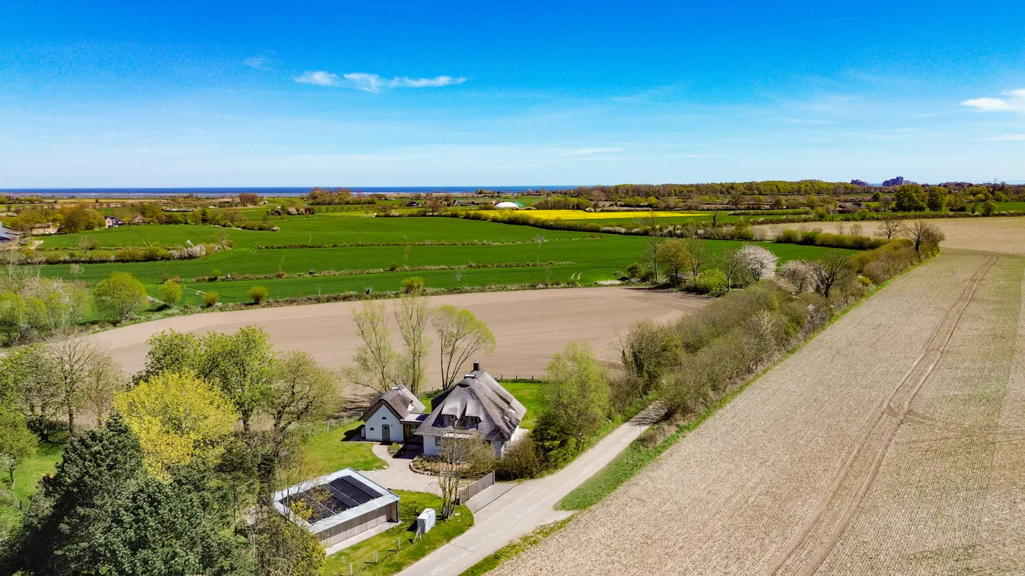 Aerial view of a thatched-roof cottage surrounded by vast green and brown agricultural fields under a clear blue sky.