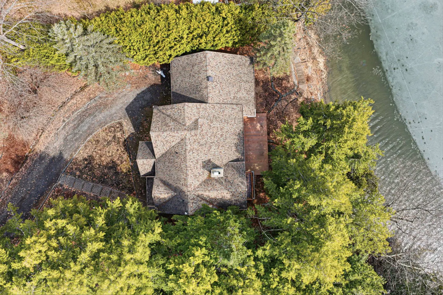 Aerial view of a brown-roofed house surrounded by green trees, a driveway, and a partially frozen lake.