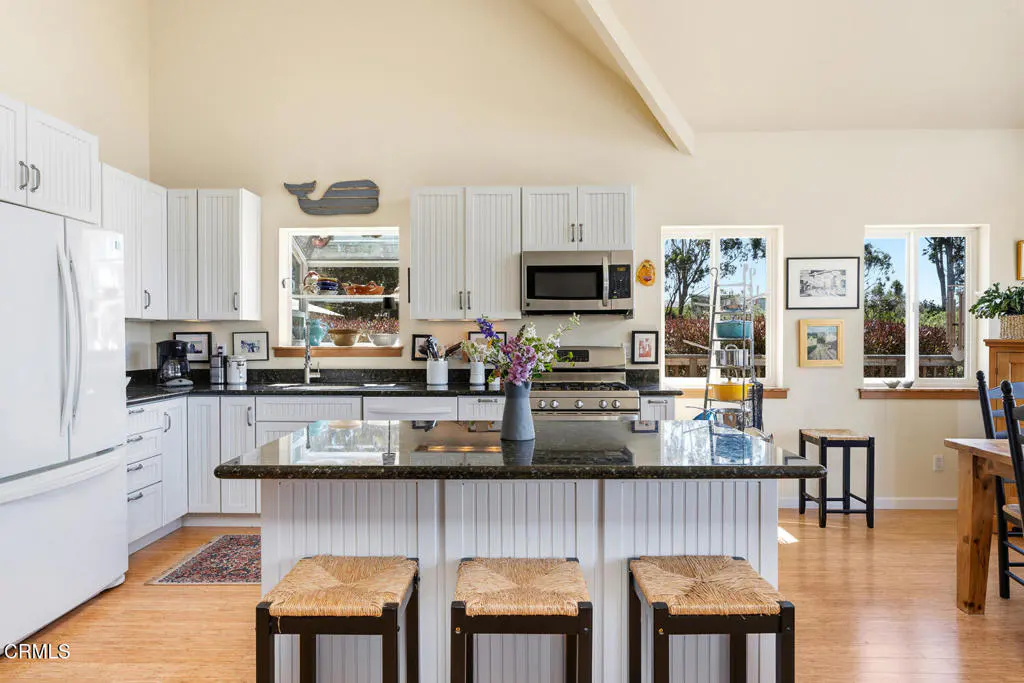 Bright kitchen with white cabinets, black countertops, and a central island with three woven stools. Windows offer views of greenery.