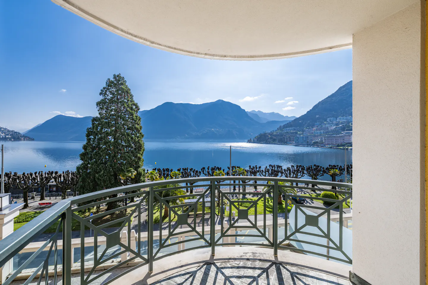 Balcony view of a blue lake and mountains under a clear sky. A green metal railing is in the foreground.