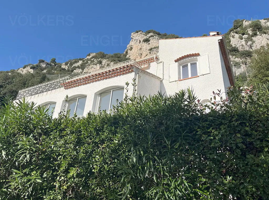 Exterior view of a white stucco house with red tile roof, partially obscured by green bushes, against a rocky hillside and blue sky.