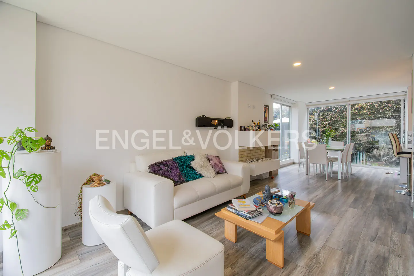 Bright living room with white sofa, wood floors, and a dining area by large windows overlooking greenery. Engel & Volkers logo visible.