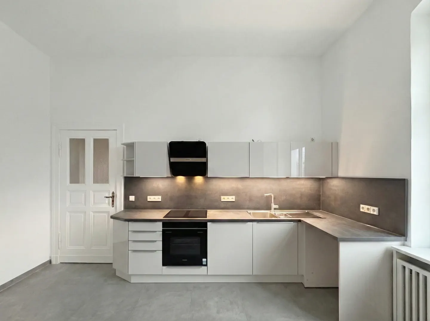 Bright, modern kitchen with white cabinets, gray countertops, and stainless steel sink. Black range hood above the stove. White door on the left.