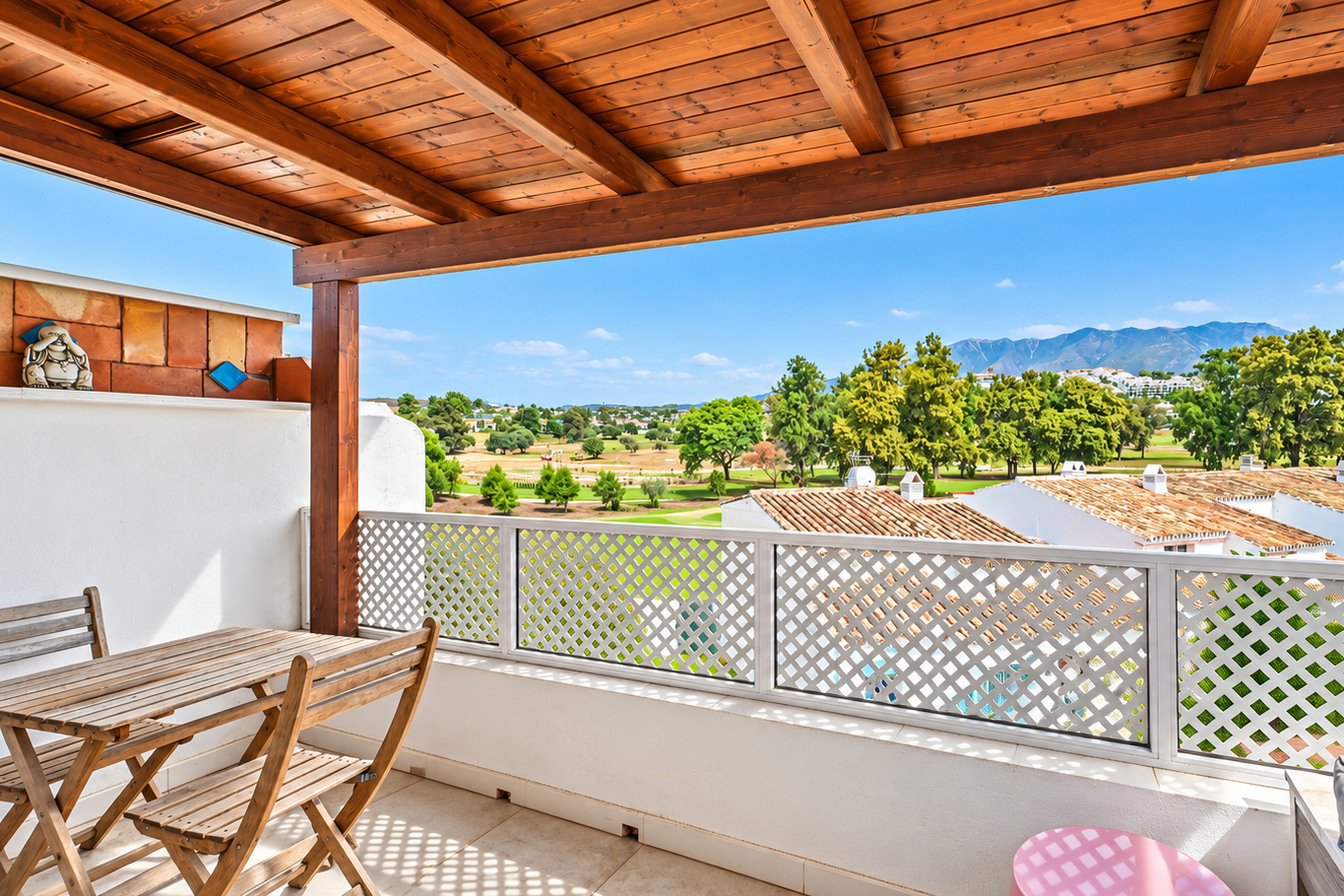 Covered balcony with wooden table and chairs, white lattice railing, and a scenic view of trees, hills, and blue sky. [AI-generated text]