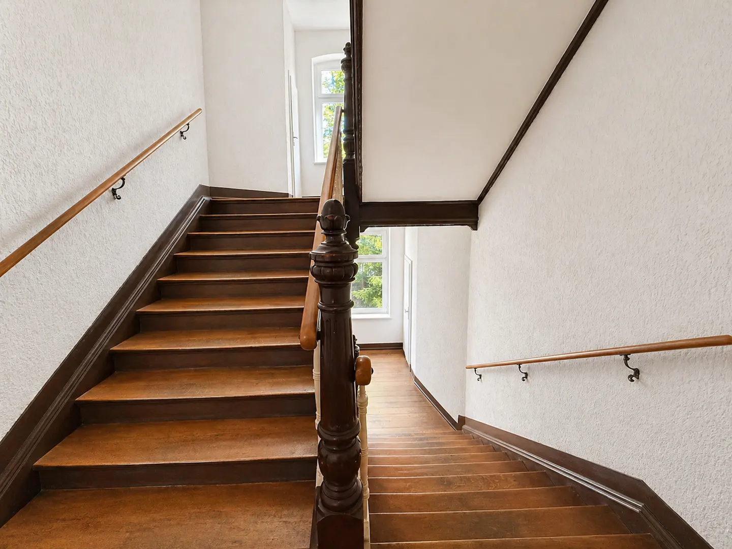 Wooden staircase with dark trim and handrails in a white-walled building, leading up and down. Windows visible in the background.