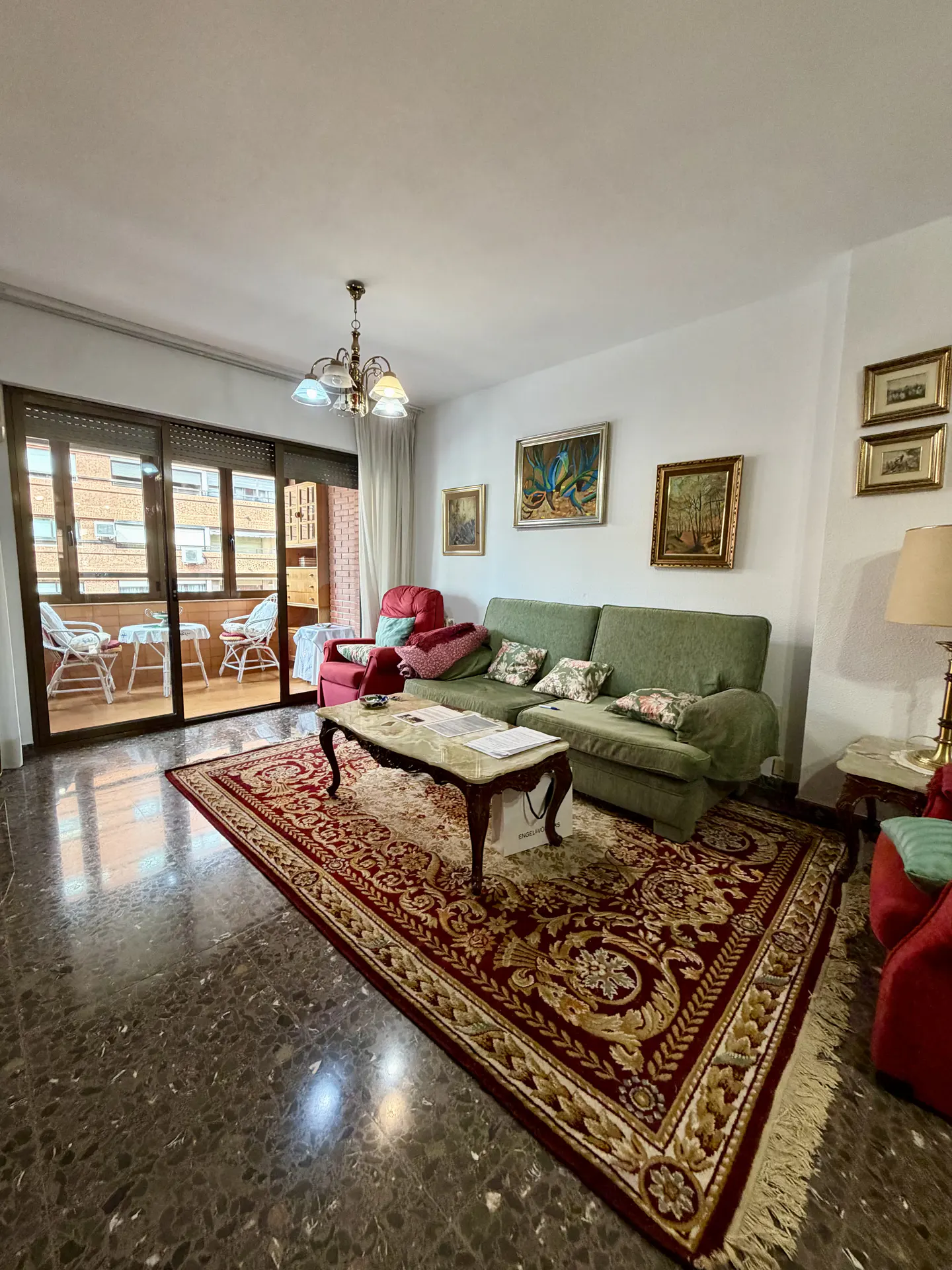 Living room with green sofa, red armchair, and patterned rug. Balcony with chairs visible through sliding glass doors.
