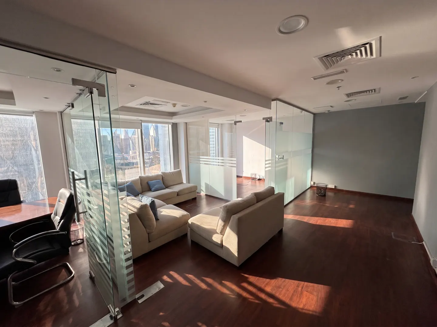 A bright office space with glass walls, tan sofas, and a dark wood floor. A desk and chair are visible in the foreground.