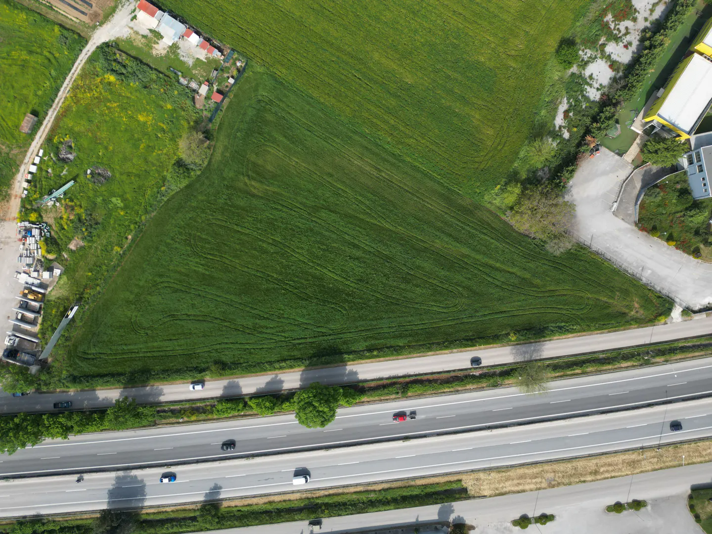 Aerial view of a green field next to a highway with cars, buildings, and trees.