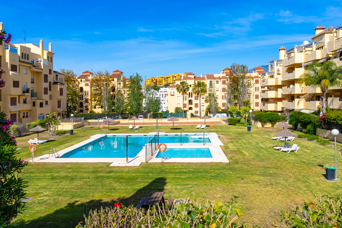 Exterior view of a complex with a pool, green lawn, and beige buildings under a blue sky.