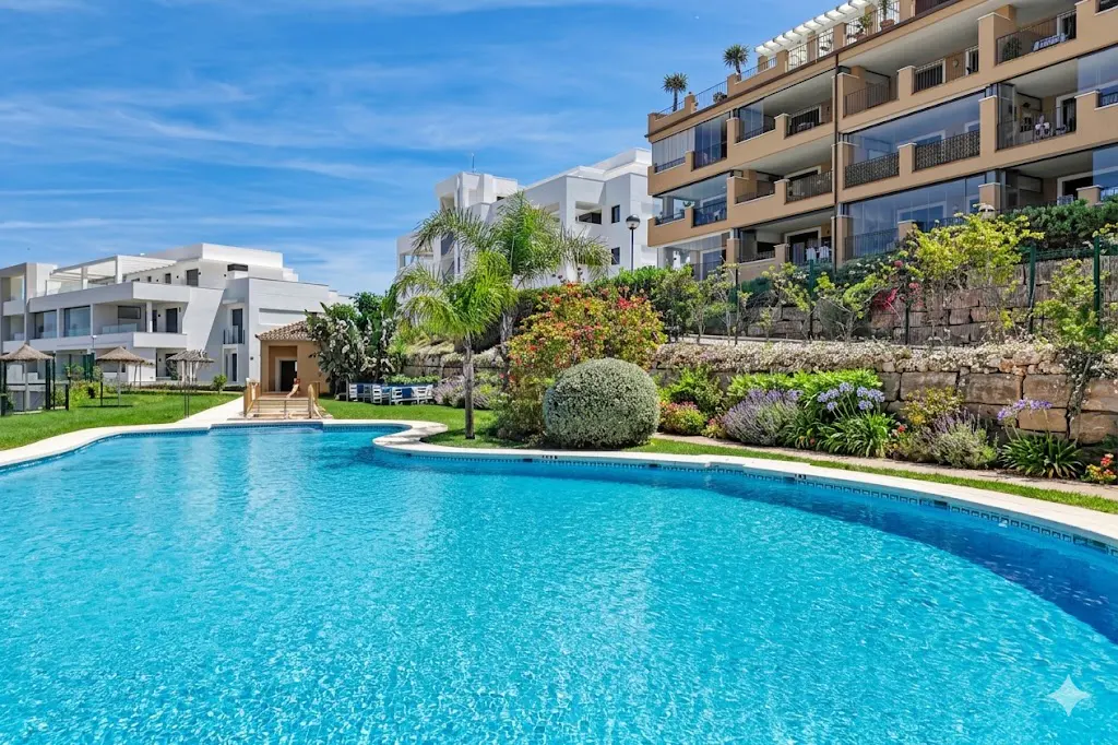 A turquoise pool sits in front of modern white and tan buildings under a blue sky with wispy clouds. Landscaping includes green grass, trees, and colorful flowers.