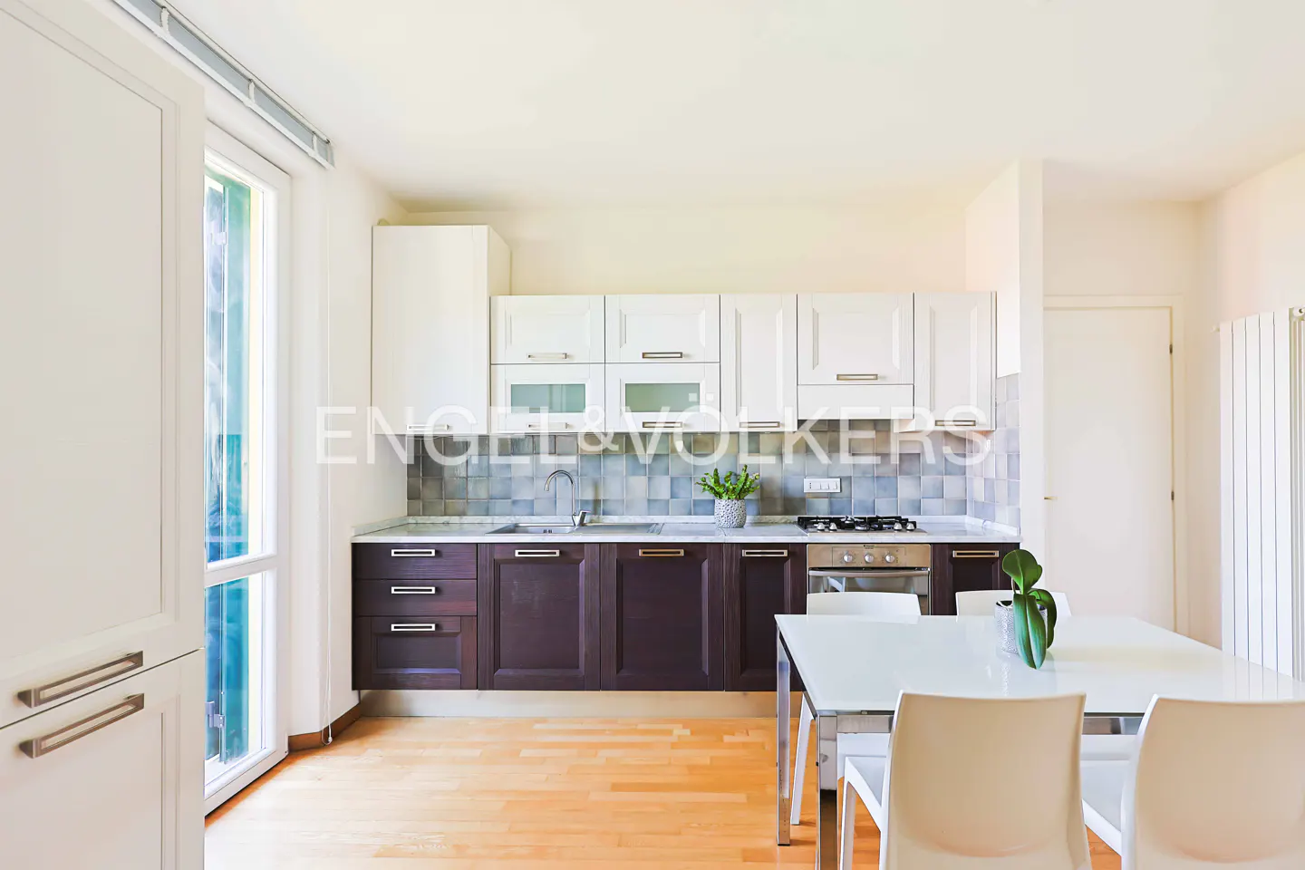 A bright kitchen with white cabinets above and dark brown cabinets below, a white table and chairs, and wood floors.