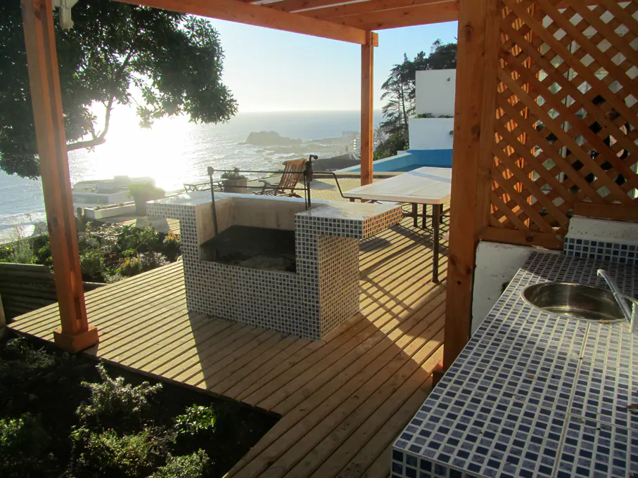 Outdoor kitchen with a tiled grill, sink, and table on a wooden deck overlooking the ocean.