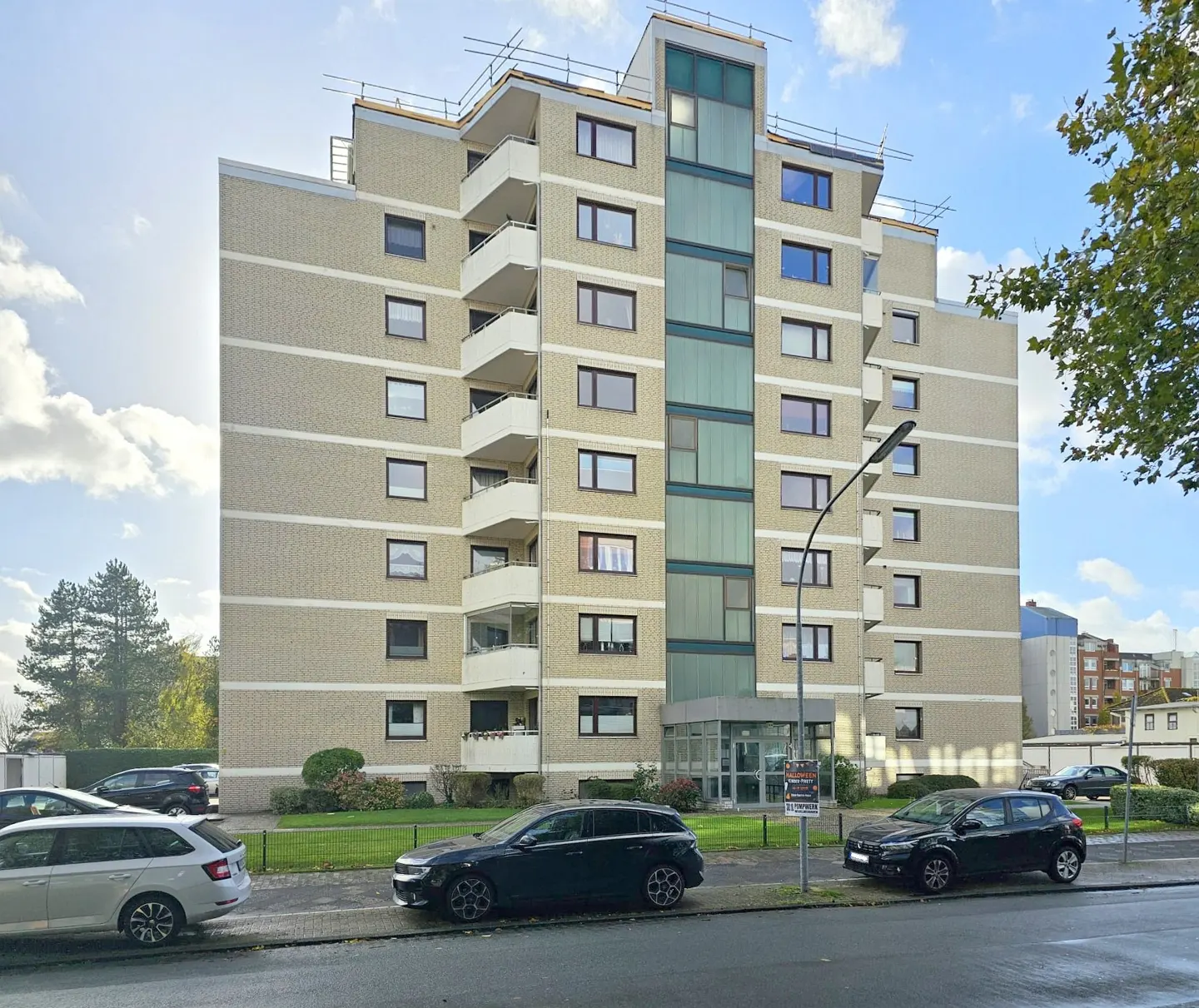 A beige apartment building with balconies and a glass elevator shaft, cars parked on the street.