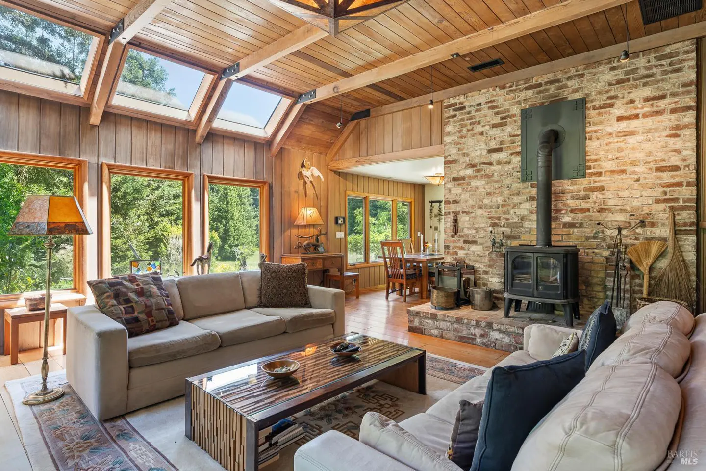 Living room with wood paneled walls, skylights, and brick fireplace. Beige sofas face a glass-topped coffee table. Windows overlook green trees.
