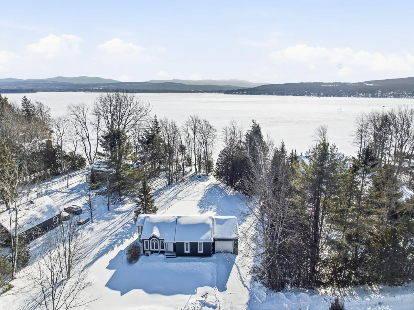 Aerial view of a dark blue house with snow-covered roof and yard, near a frozen lake and trees under a blue sky.
