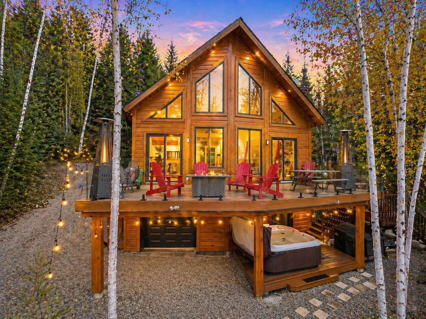 Wooden cabin with a deck, red chairs, and a hot tub, surrounded by trees and string lights at dusk.