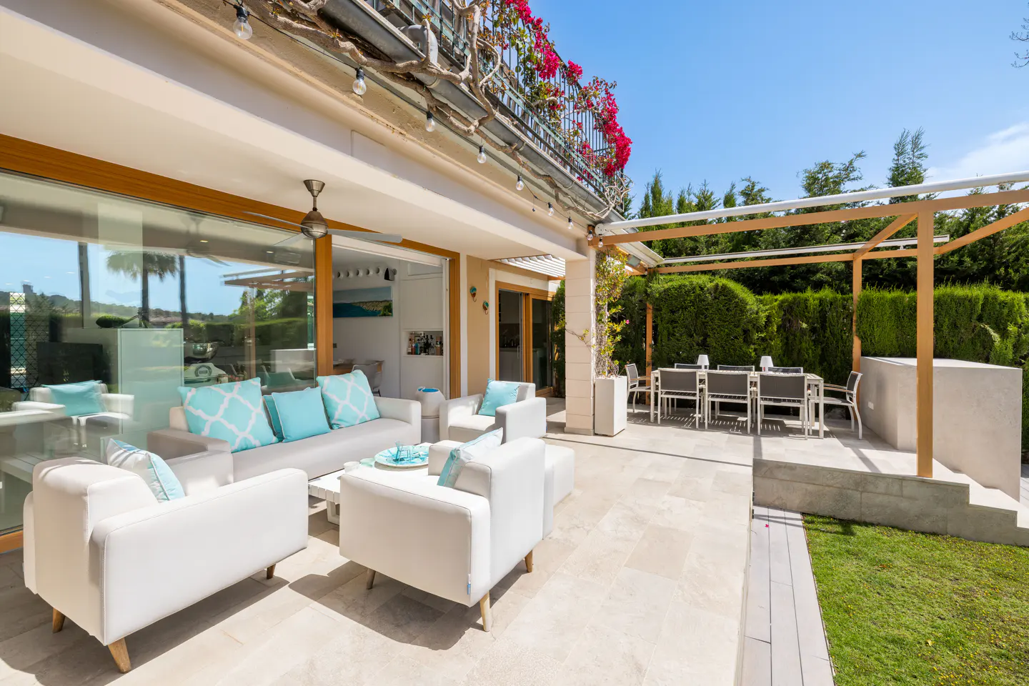 Outdoor patio with white furniture, turquoise pillows, and a dining table under a pergola. Red flowers adorn the balcony above.