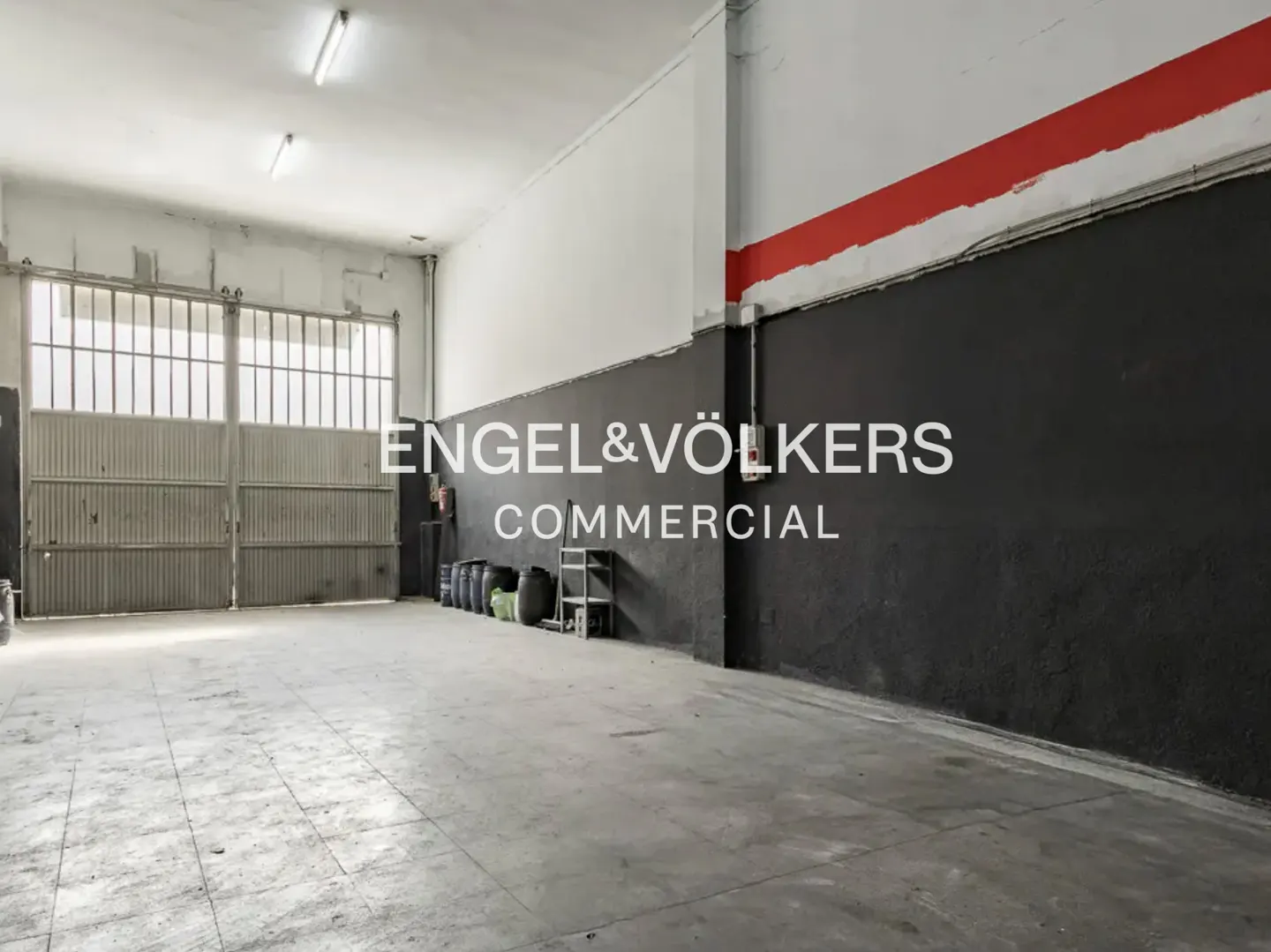 A large, empty commercial space with gray concrete floors, a black and white wall with a red stripe, and a metal gate.