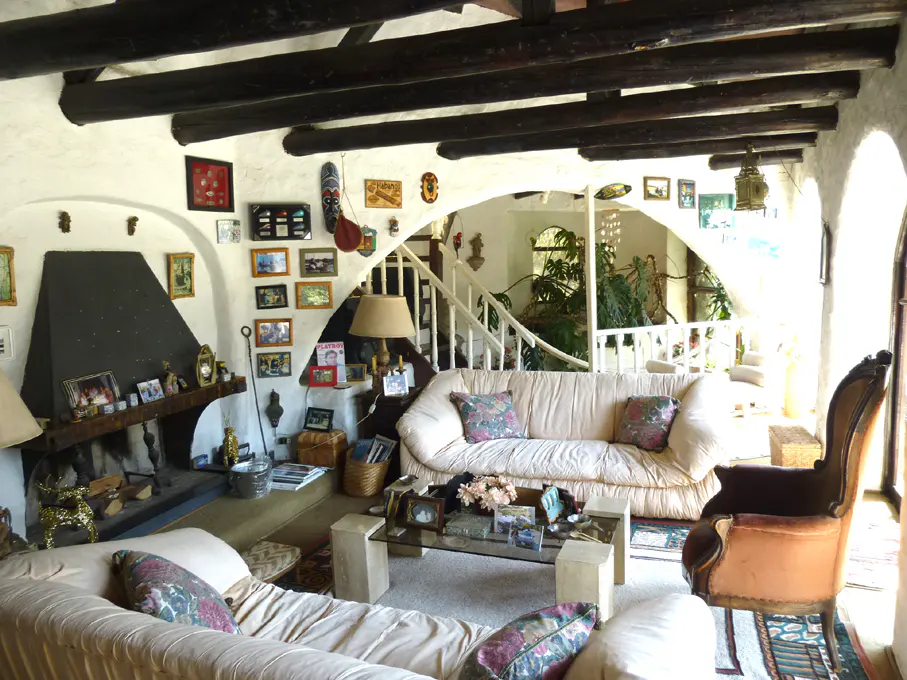 Living room with white walls, dark wood beams, and a black fireplace. Two sofas, a glass coffee table, and a staircase are visible.