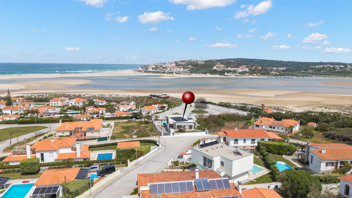 Aerial view of a modern white house with a red pin, near a beach and town under a blue sky.