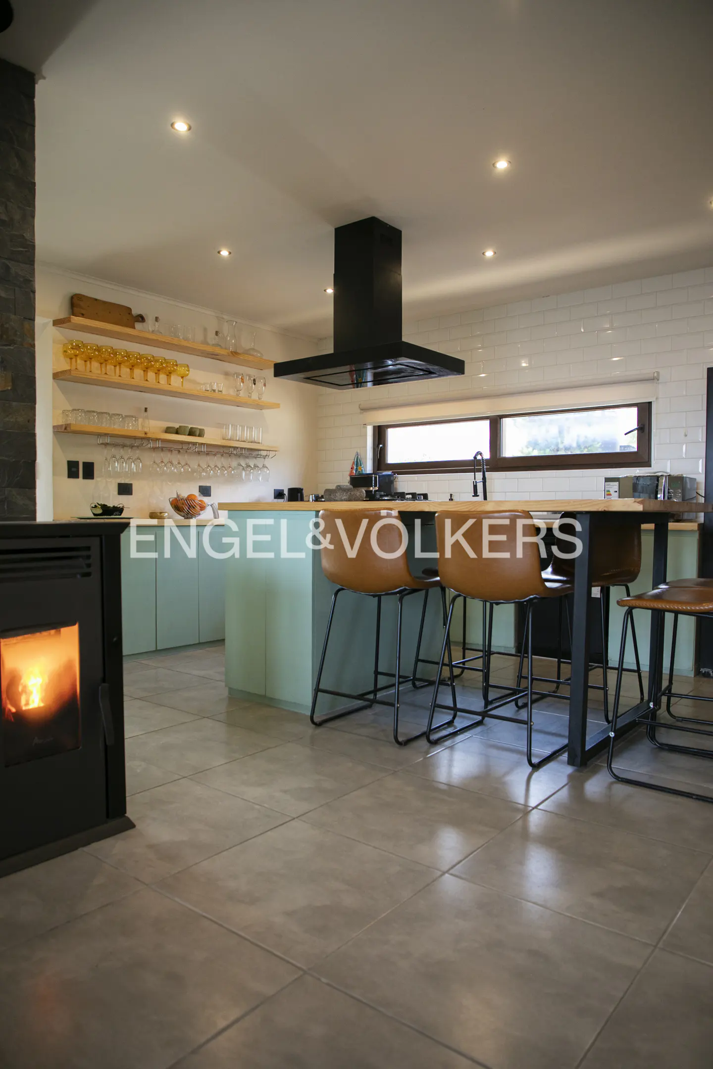 Modern kitchen with light green island, tan leather bar stools, black range hood, and a cozy fireplace in the foreground.