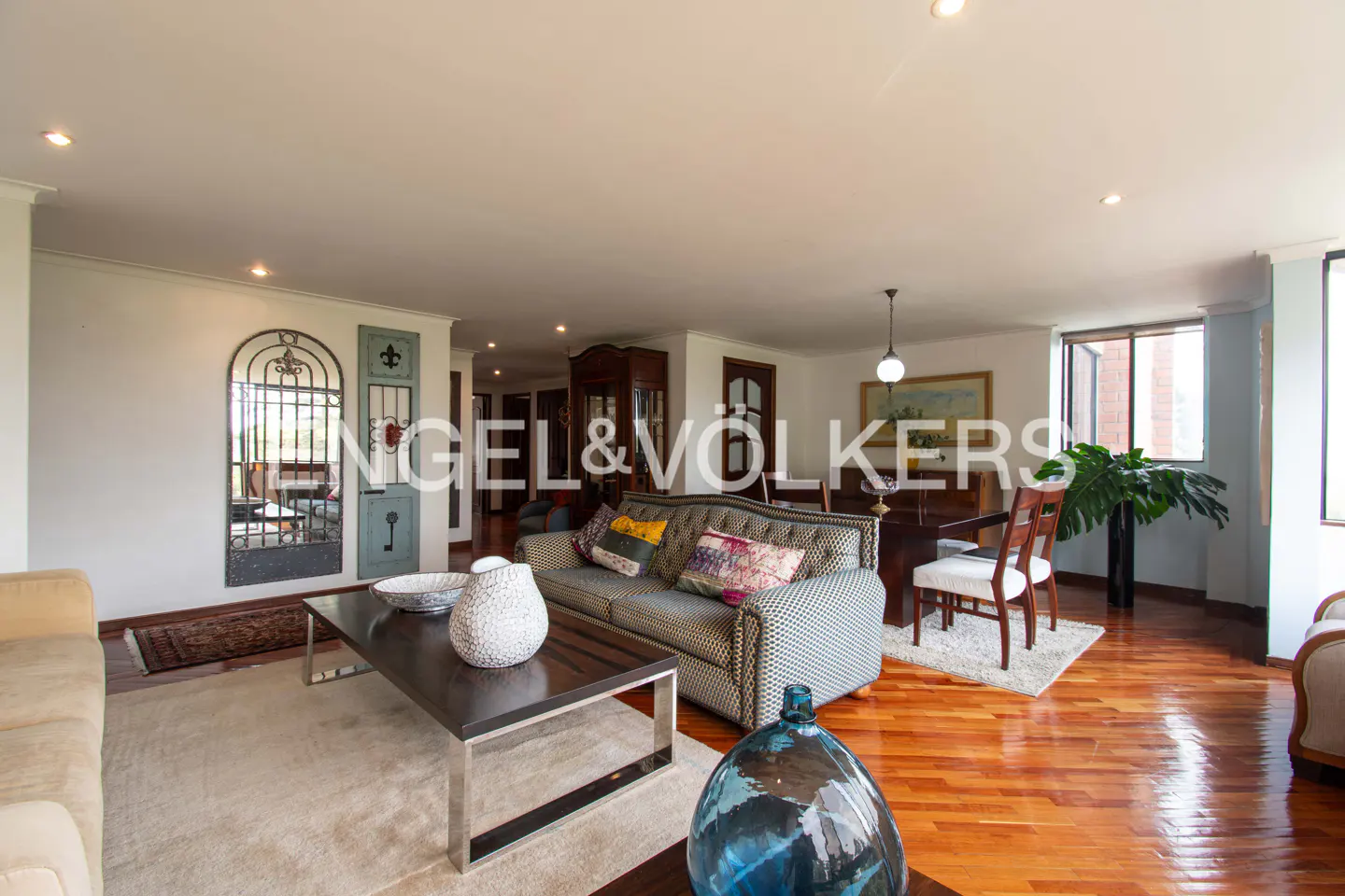 Spacious living room with hardwood floors, patterned sofa, dark coffee table, dining area, and decorative arched mirror.