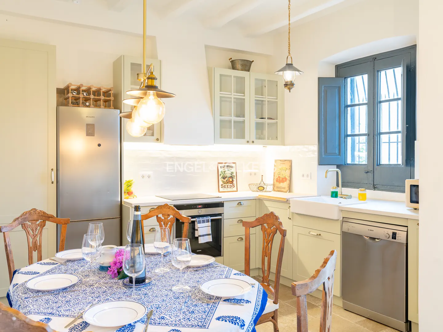 Bright kitchen with a round table, blue patterned tablecloth, wooden chairs, stainless appliances, and a farmhouse sink.