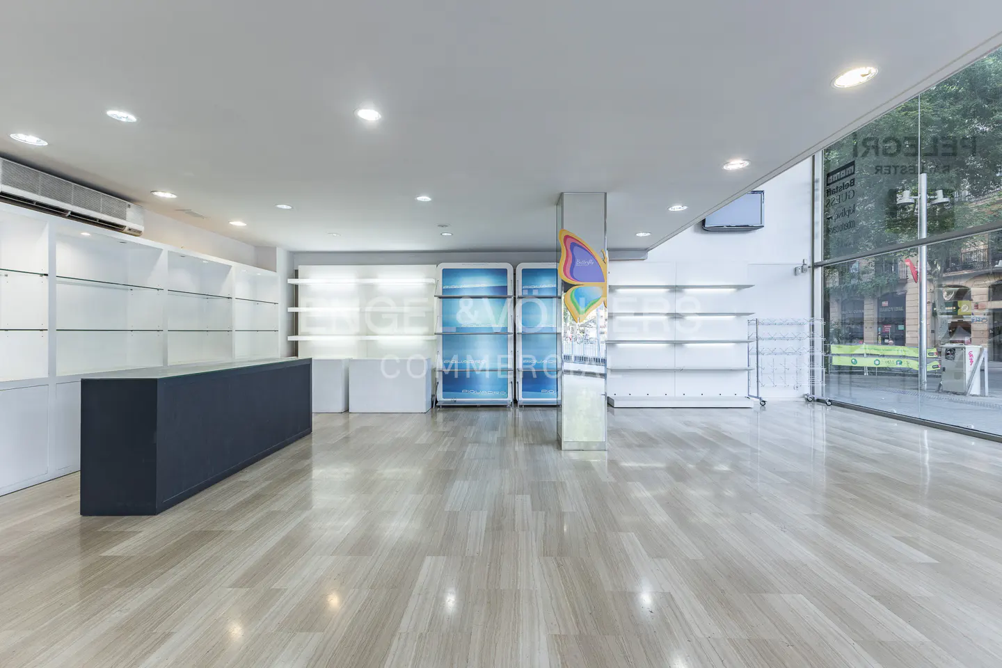 Bright, empty retail space with light wood floors, white walls, and large windows. A dark gray counter sits to the left.