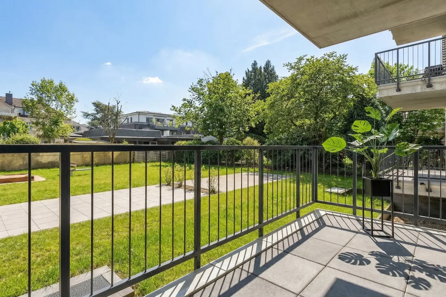 Balcony view of a green lawn with trees, seen through a black metal railing. A potted plant sits on the gray tiled balcony.