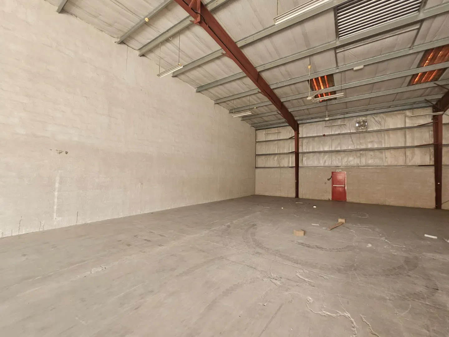 Empty warehouse interior with concrete floor, metal roof beams, and a red door in the back wall.
