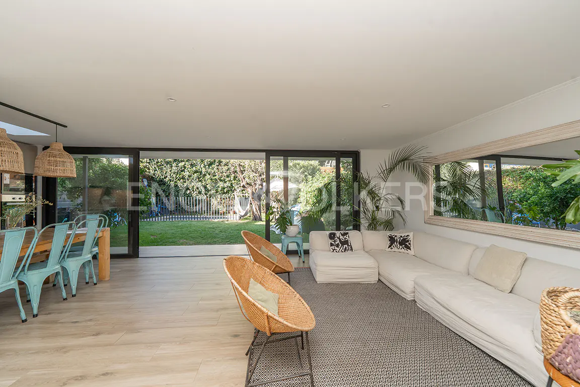 Bright, open living space with a white sofa, wicker chairs, and a dining table with blue chairs. Large windows overlook a green lawn.