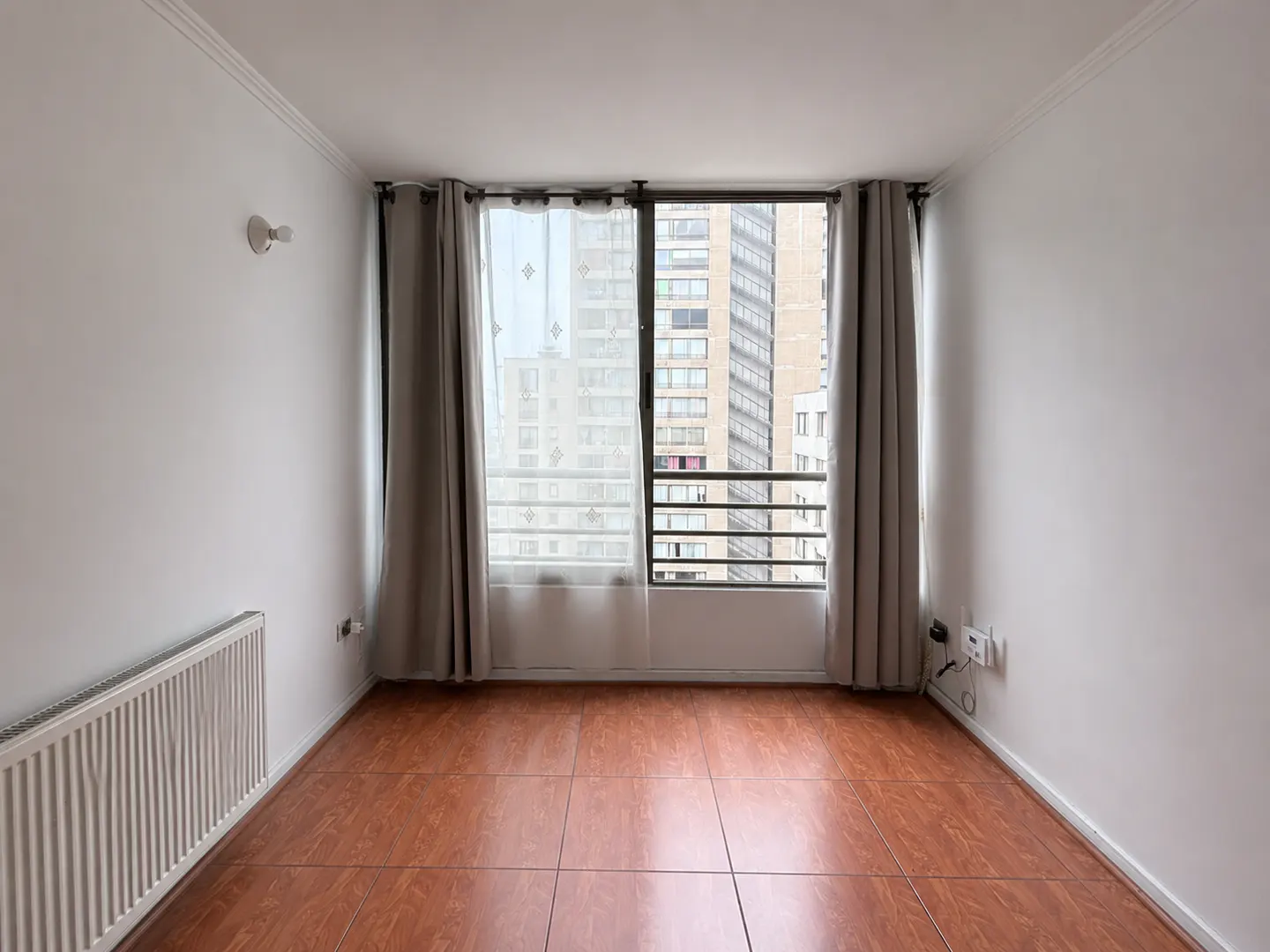 Empty room with white walls, brown tile floor, and a window with gray curtains overlooking city buildings.