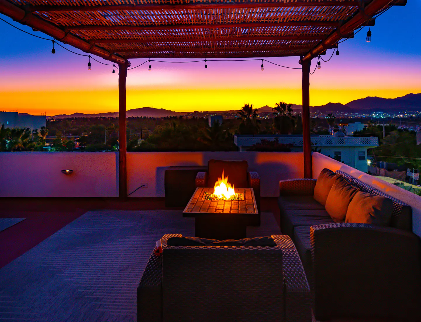 Rooftop patio with wicker furniture, fire pit, and sunset view. String lights hang from a bamboo pergola.
