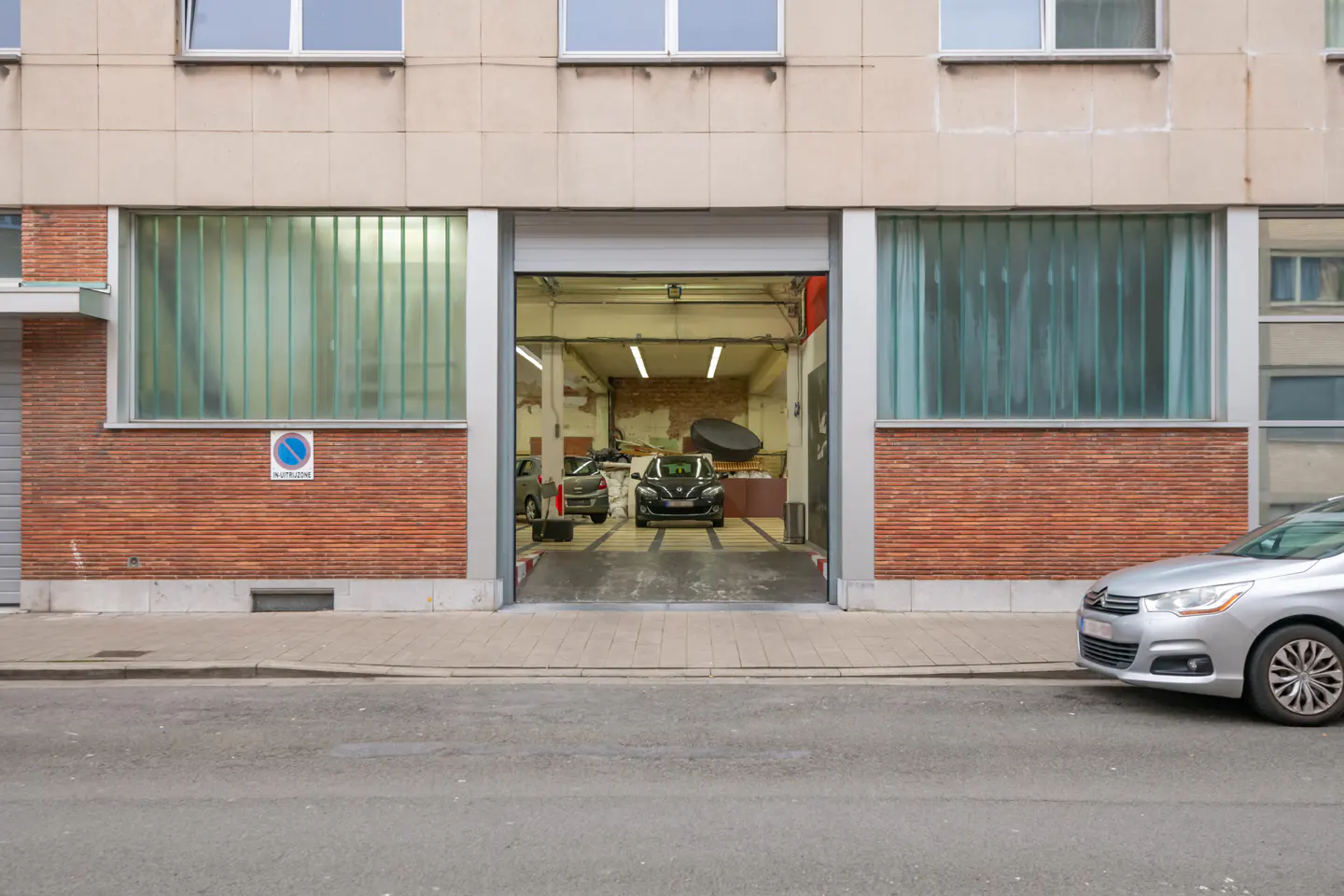 Exterior of a brick building with a garage door open, revealing two cars inside. A silver car is parked on the street.