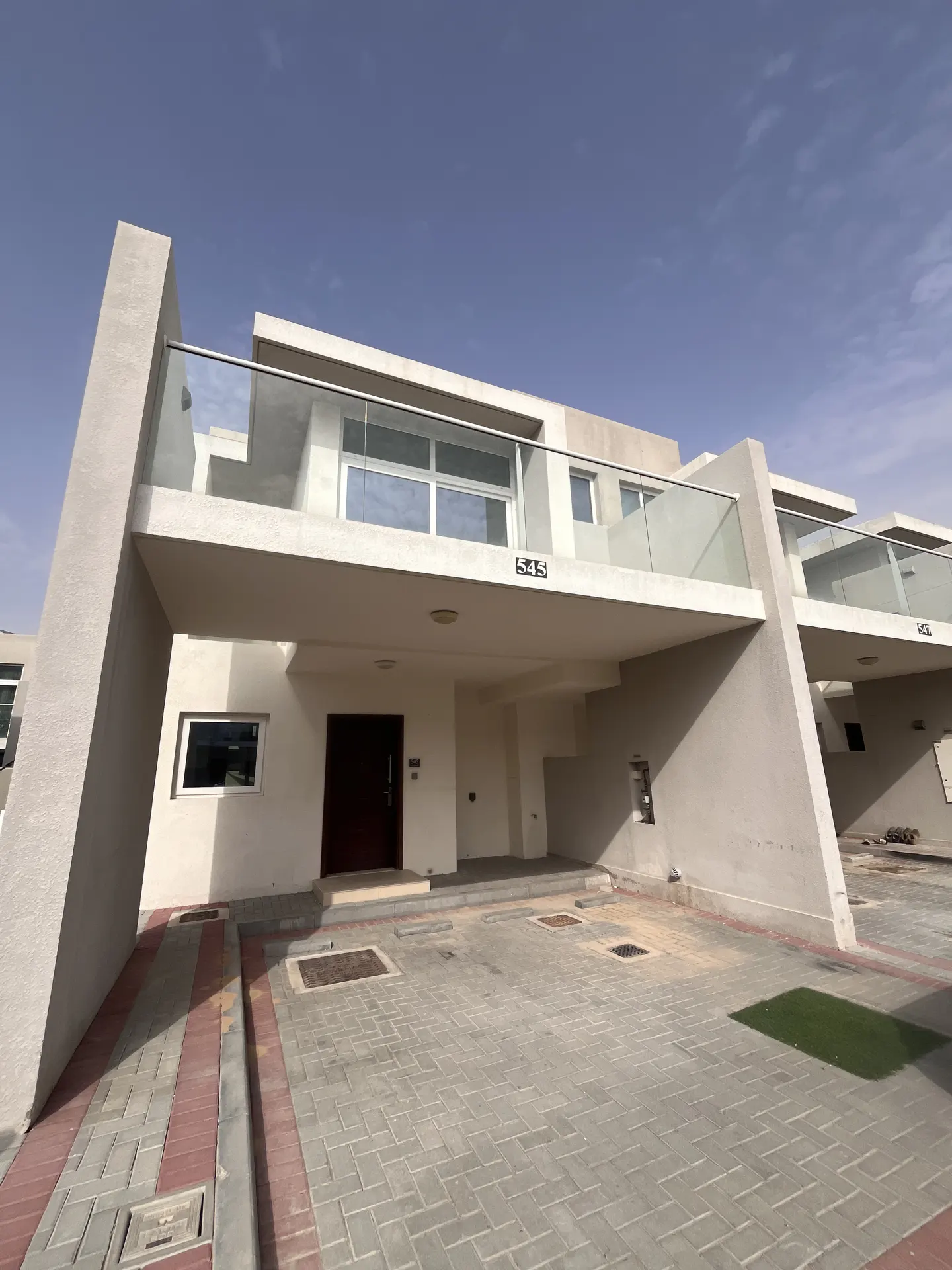 Modern two-story townhouse with a beige exterior, glass balcony, and a dark brown front door. Paved driveway in front.