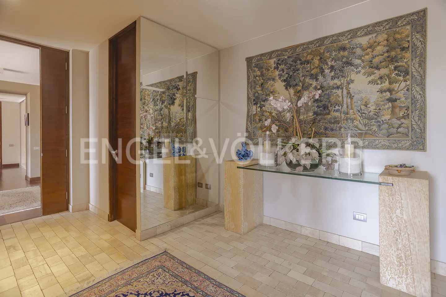 Hallway with stone tile floor, a glass console table, a large landscape tapestry, and a mirrored wall with a wooden door.