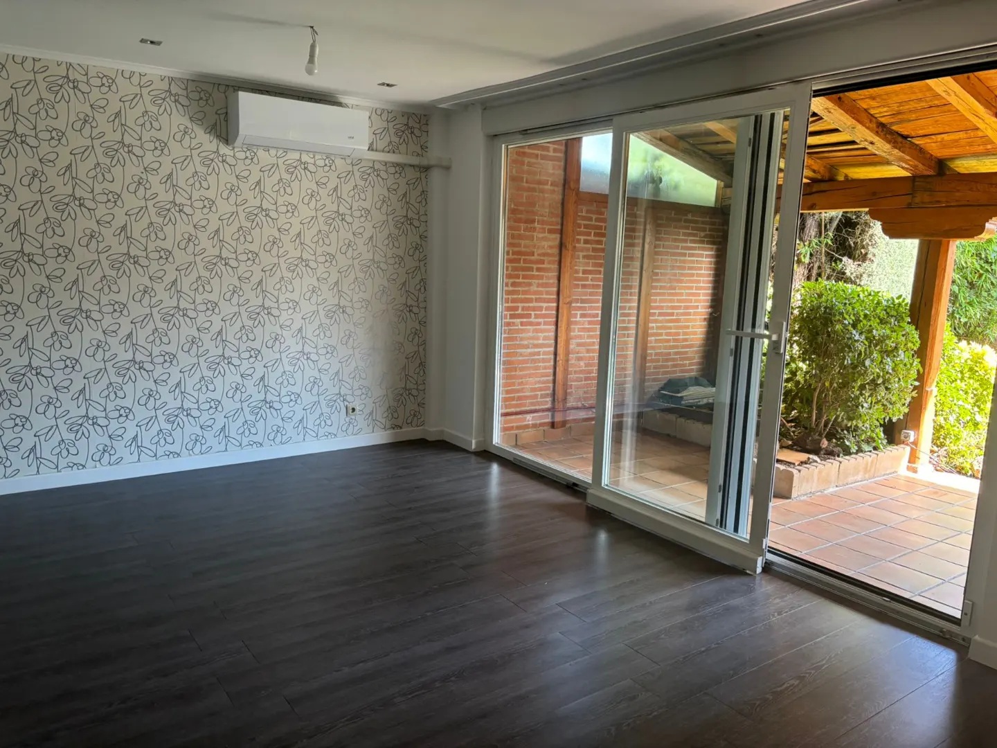 Living room with dark wood floors, floral wallpaper, air conditioning unit, and sliding glass doors to a brick patio.