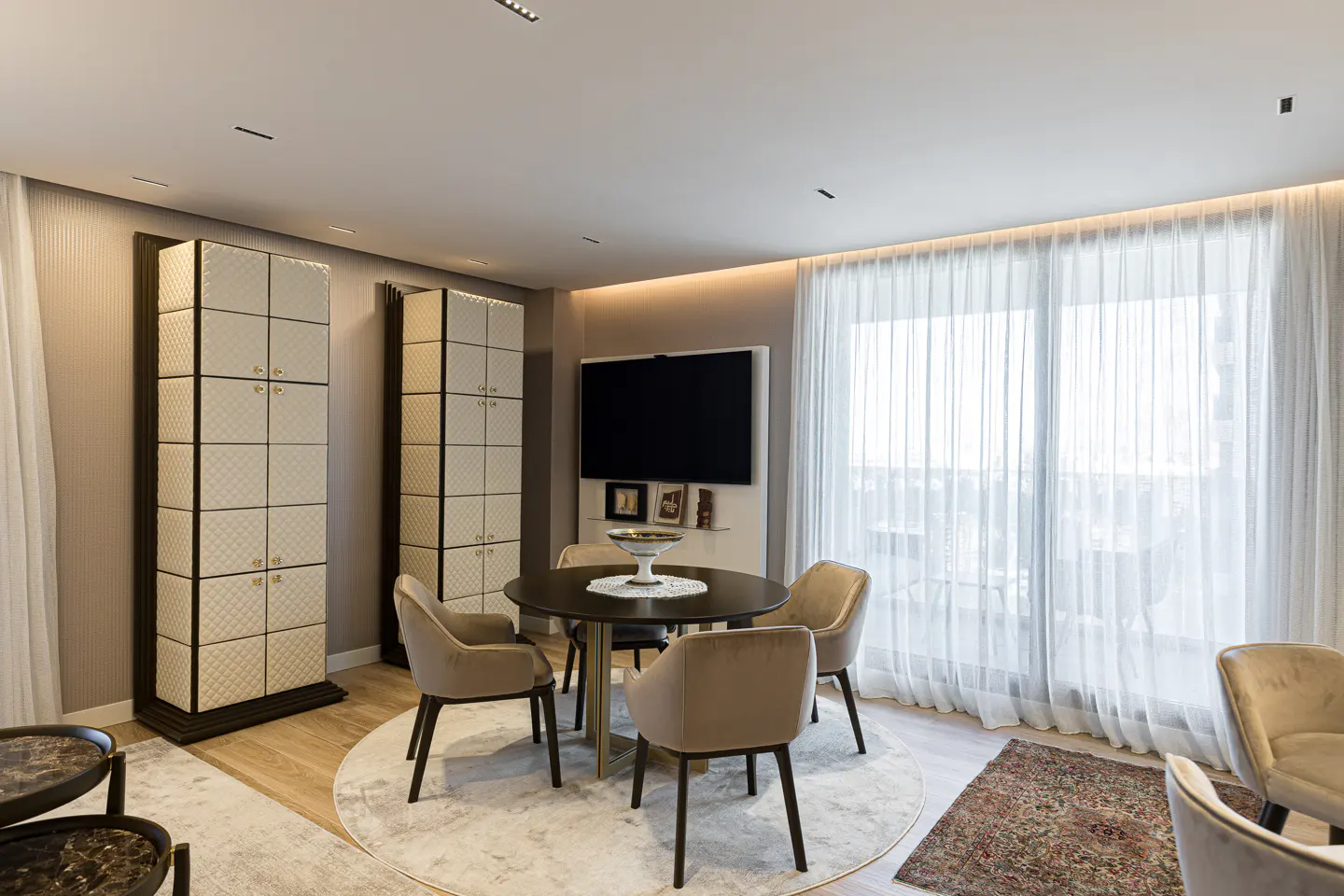 Living room with a round table, four beige chairs, a TV, and two tall cabinets with patterned doors. White curtains cover a large window.
