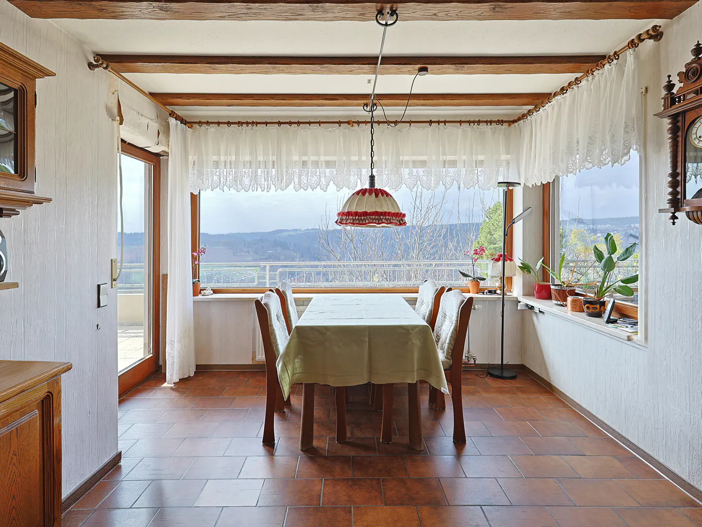 Dining room with a table covered in a light green cloth, surrounded by wooden chairs. Large windows offer a view of a green landscape. The floor is tiled in brown.