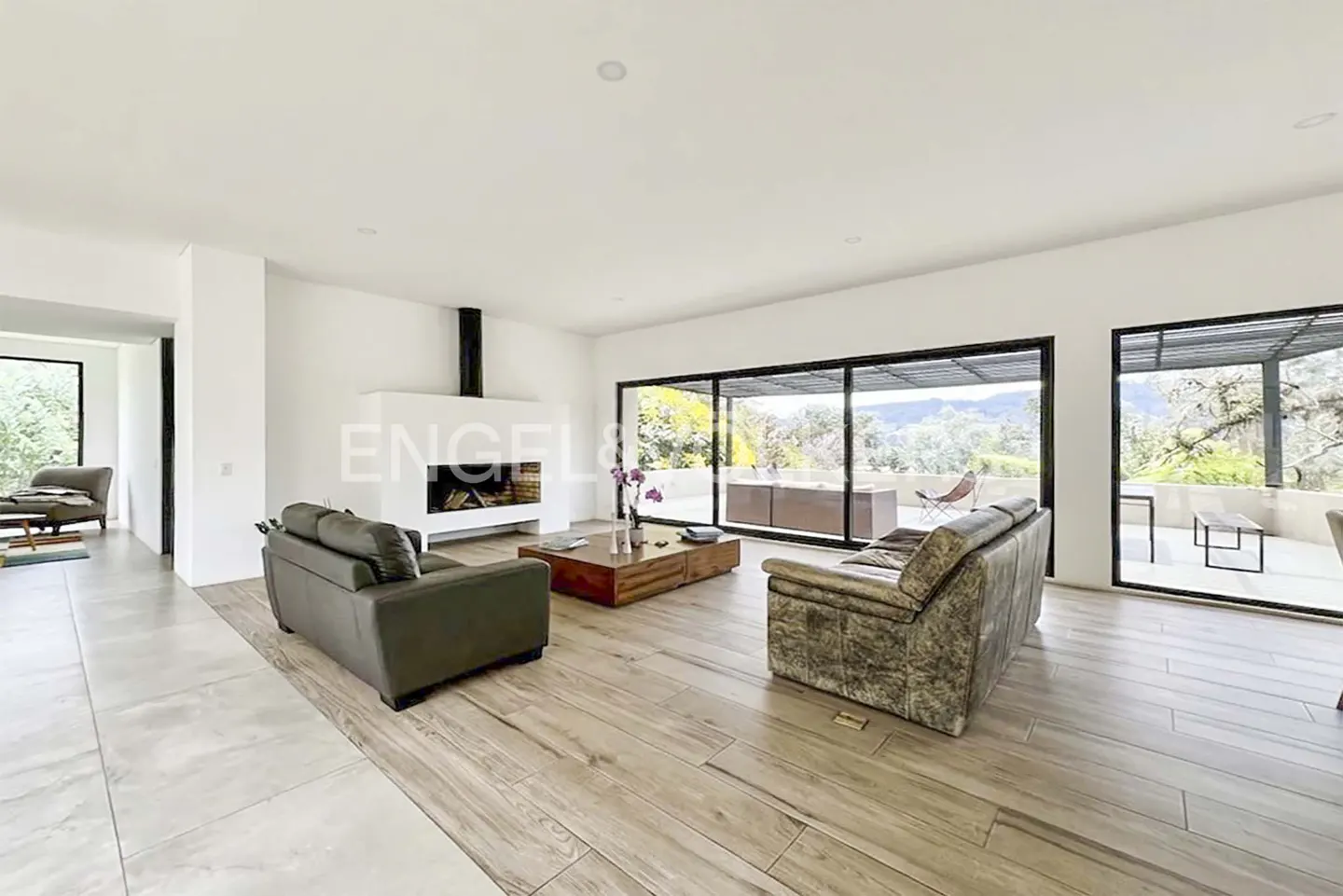 Bright living room with wood floors, white walls, and large windows. Two sofas face a modern fireplace and a wood coffee table with flowers.