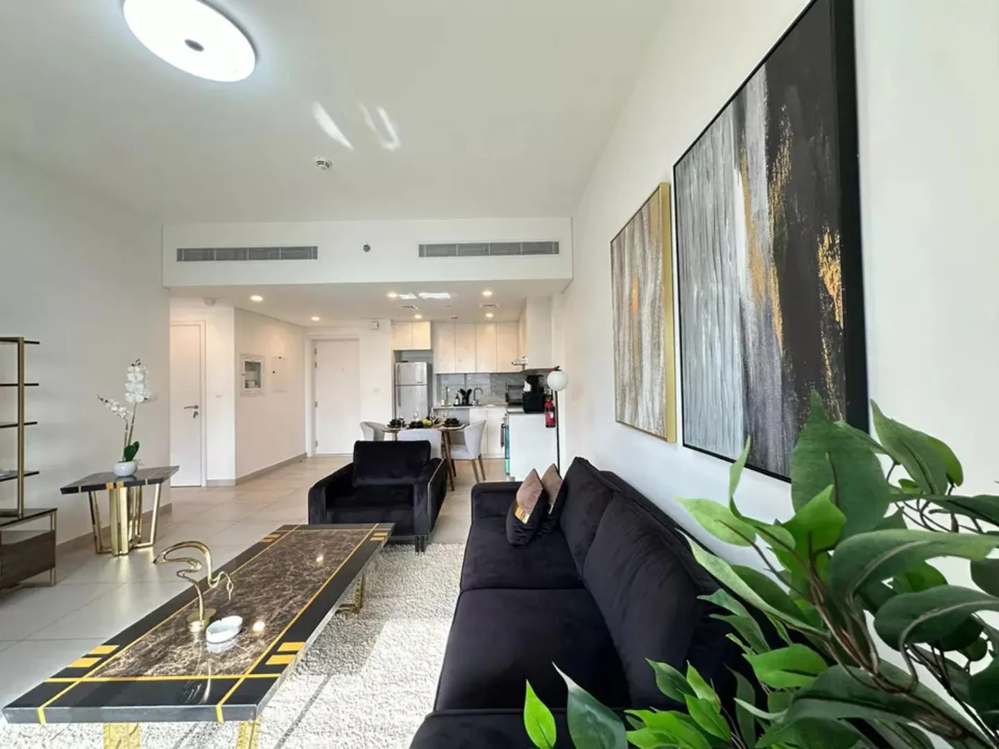 Bright living room with black velvet sofa and chair, marble coffee table, and abstract art on white walls. Kitchen visible in the background.
